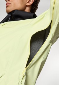 Veste jaune clair avec un col noir, dotée d'une poche zippée qui révèle une doublure intérieure gris foncé. Tissu lisse et léger.