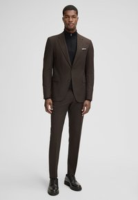 Brauner Anzug mit einem Revers mit Kerbe, kombiniert mit einem schwarzen Zip-Up-Oberteil. Der Anzug hat eine maßgeschneiderte Passform und wird durch schwarze Schuhe ergänzt.