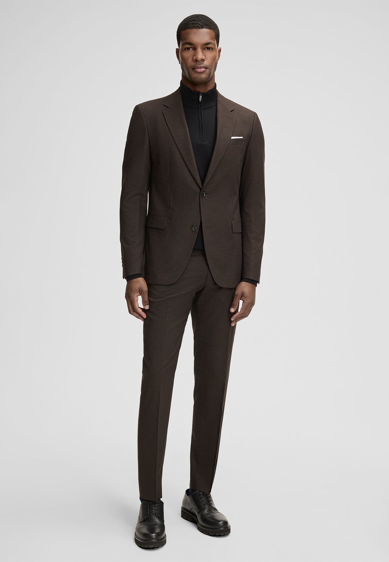 Brauner Anzug mit einem Revers mit Kerbe, kombiniert mit einem schwarzen Zip-Up-Oberteil. Der Anzug hat eine maßgeschneiderte Passform und wird durch schwarze Schuhe ergänzt.