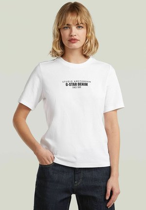 Jeune femme aux cheveux blonds portant un t-shirt blanc G-Star Denim et un jean foncé, debout avec une main dans la poche.