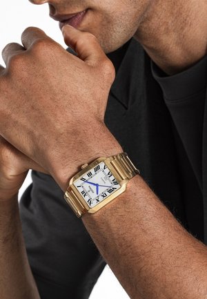Montre rectangulaire en or avec un cadran blanc à chiffres romains et des aiguilles bleues. Bracelet en métal à maillons avec finition polie. Portée sur un bras marron.