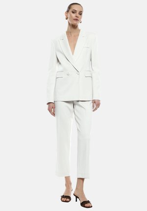 Blazer bianco su misura con doppi bottoni, scollatura a V profonda, abbinato a pantaloni bianchi con risvolto. Il modello indossa sandali neri con tacco e gioielli semplici.