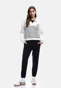 Hvid zip-up sweatshirt med sorte striber, der har et logo, parret med sorte joggers og hvide sneakers med sorte accenter.