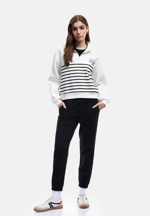Witte zip-up sweatshirt met zwarte strepen, met een logo, gecombineerd met zwarte joggers en witte sneakers met zwarte accenten.