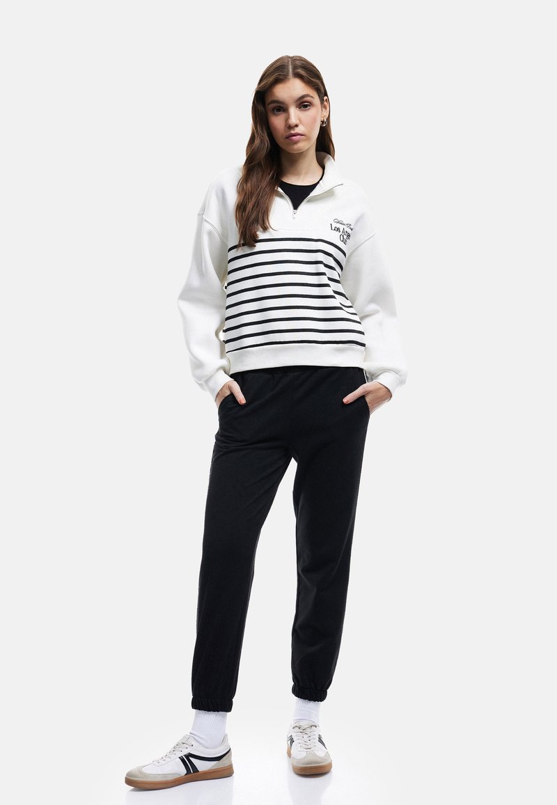Hvid zip-up sweatshirt med sorte striber, der har et logo, parret med sorte joggers og hvide sneakers med sorte accenter.
