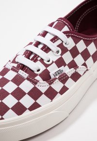 Baskets Vans à carreaux rouge marron et blanc avec lacets blancs, œillets en métal et semelle en caoutchouc blanche sur fond blanc.