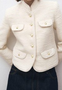 Chaqueta texturizada marfil con cuello mandarín, seis botones dorados y dos bolsillos con solapa en la parte delantera. Combinada con jeans de mezclilla oscura.