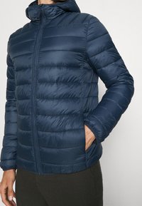 Doudoune marine avec fermeture éclair à l'avant, capuche, design matelassé et poches latérales. Fabriquée en matériau léger et résistant à l'eau.