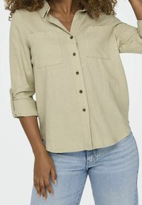 Lys beige button-up skjorte med lange ærmer, brystlommer og en afslappet pasform; lavet af tekstureret stof, båret over blå jeans.