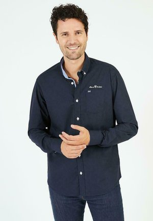 Chemise bleu marine à boutons avec un col bleu accentué, une poche poitrine et un logo brodé. Tissu lisse avec une coupe ajustée.