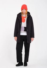 Sort polstret jakke med hætte, båret over en flerfarvet sweater. Kombineret med løse sorte bukser og en klar rød hue.