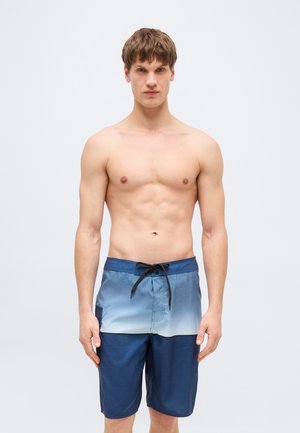 Νεαρός άνδρας που στέκεται προς τα εμπρός φορώντας μπλε σορτς κολύμβησης με μαύρο κορδόνι, χωρίς μπλούζα, ουδέτερο φόντο.