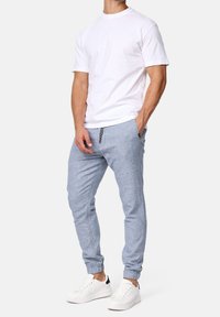 Man wearing plain white short-sleeve t-shirt, light blue jogger pants with drawstring, and white sneakers, standing with one hand in pocket.