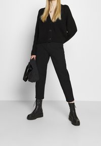 Černý pletený kardigan s knoflíky, spárovaný s černými zúženými kalhotami a černými šněrovacími bojovými botami. Oblečení je monochromatické a minimalistické.