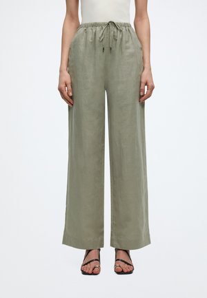 Kvinne iført olivengrønne vid bukser i lin med elastisk snor i livet og svarte sandaler med stropper og hæl, stående mot en ensfarget bakgrunn.