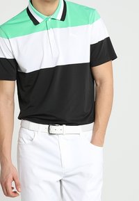 Polo à col avec rayures horizontales vertes, blanches et noires. Tissu léger, boutons contrastants, associé à une ceinture blanche et un pantalon blanc.
