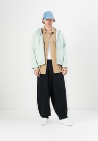 Light green hoodie over a tan button-up shirt, paired with black wide-leg trousers and white sneakers. Blue cap completes the outfit.