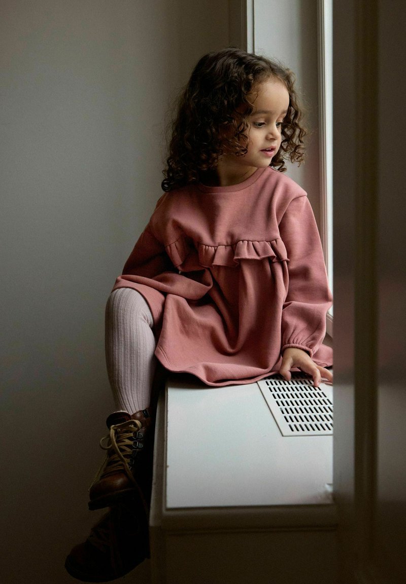 Jeune fille aux cheveux bouclés, vêtue d'une robe rose et de collants, assise sur un cache-radiateur, regardant par une fenêtre proche avec sa main posée sur la ventilation.
