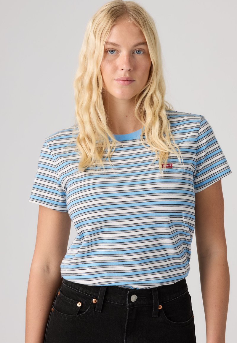Femme blonde aux cheveux ondulés portant un t-shirt rayé bleu et blanc et un jean noir taille haute, debout devant un fond uni.