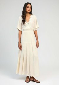 Robe longue couleur crème avec un décolleté en V profond, des manches courtes à volants et une jupe à étages. Détails en subtitres le long de l'encolure.