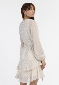 Rochie albă, stratificată, cu mâneci lungi, transparente; prezintă broderie florală aurie, talie strânsă și un tiv volanat. Material fin, ușor.