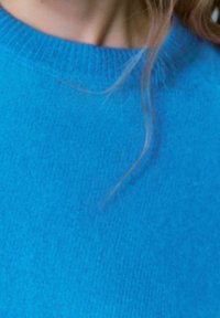 Pullover lavorato a maglia di un blu intenso con scollo tondo e texture morbida. Ciocche di capelli visibili incorniciano parzialmente l'area del colletto.