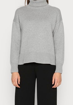 Kobieta w jasnoszarym, prążkowanym golfie i luźnych czarnych spodniach, stojąca z ramionami swobodnie opadającymi wzdłuż ciała.