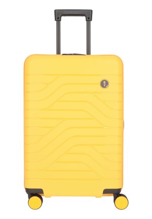 Gelber Hartschalenkoffer mit gerilltem Design, ausgestattet mit einem verstellbaren Griff, vier schwarzen Rädern und einem runden Logo auf der Vorderseite.