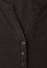 Detailní záběr na tmavě hnědou košili s výstřihem do V, se třemi svisle uspořádanými černými knoflíky a viditelnými švy.