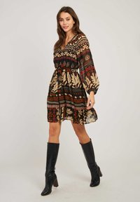 Femme marchant dans une robe à motifs arrivant au genou avec des manches longues et des bottes noires à talons hautes contre un fond uni.