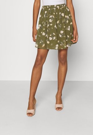 Femme portant une jupe mi-longue à motif floral vert olive et des sandales beiges à talons blocs et orteils ouverts.