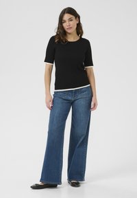 Zwarte ribbelige top met korte mouwen en crème bies, gecombineerd met blauwe wijde denim jeans. Model staat op een witte achtergrond.