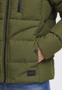 Jachetă verde căptușită, cu închidere prin fermoar, secțiuni mari matlasate și un nasture de prindere din metal negru. Include o etichetă cu logo pe lateral.