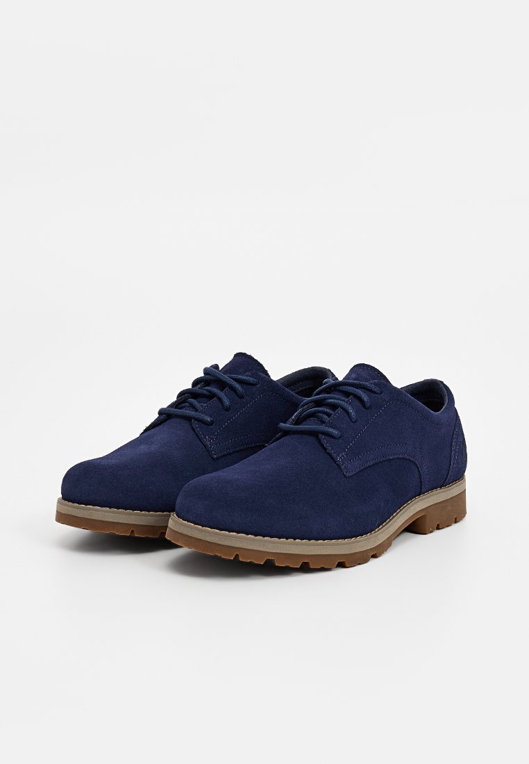 Scarpe stringate in suede blu navy con suole in gomma marrone, punte arrotondate e tacchi bassi, mostrate affiancate su uno sfondo bianco.