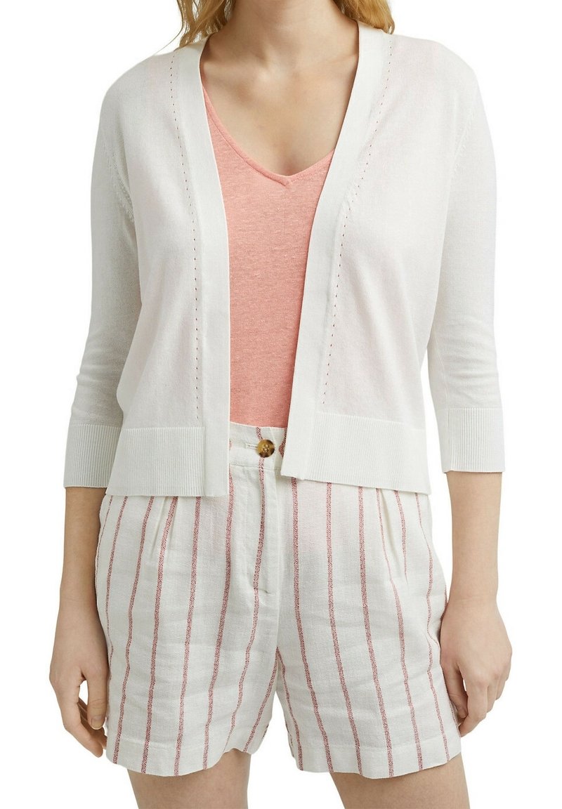 Valkoinen neuletakki, jossa on 3/4-hihat, avoin etuosa ja hienovaraiset ommelluskoristeet. Puettu vaaleanpunaisten toppien ja vaaleanraidallisten shortsien päälle.