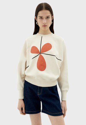Sweatshirt crème avec un design abstrait rouge et noir, col et poignets côtelés, associé à un short en denim bleu foncé. Texture lisse.