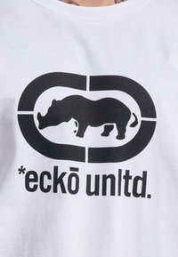 Balta marškinėliai su dideliu juodu stilizuoto rino logotipu, esančiu ovalo formos. Po logotipu užrašyta "*eckō unltd."