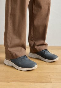 Des chaussures en suede gris avec des semelles en caoutchouc blanches, un design à lacets et de légères perforations. Portées avec un pantalon en tissu brun sur une surface en bois.