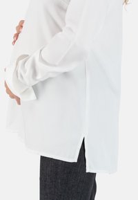 Witte blouse met een ontspannen pasvorm en zijsplitten, gemaakt van een soepele stof. Heeft lange mouwen en een ronde halslijn, gecombineerd met donkere jeans.