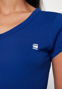 Kvinna iklädd en blå kortärmad V-ringad skjorta med en liten vit "RAW"-logotyp på vänster bröst.