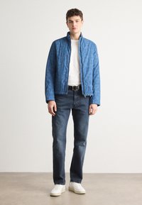 Casaco azul claro acolchoado com colarinho alto, combinado com uma t-shirt branca e jeans de ganga escura. O modelo usa ténis brancos.