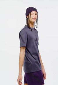 Mladý človek s vrkočmi, oblečený v purpurovej čiapke, tmavosivej tričku a purpurových šortkách, stojaci pred jednoduchým bielym pozadím.