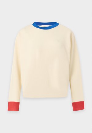 Femme portant un pull beige avec un col bleu et des poignets rouges, une jupe noire, des baskets rouges, et tenant un sac à main marron, debout sur fond blanc.