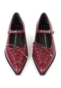 Zapatos Mary Jane de cuero rojo con punta afilada, con cierre de cordones en la parte delantera, correa con hebilla plateada y suelas negras. Textura suave.