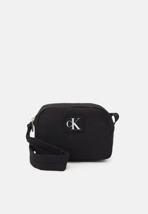 Musta suorakulmainen rannelaukku vetoketjulla suljettuna ja edessä oleva kirjailtu logo, jossa lukee "cK" valkoisin kirjaimin.