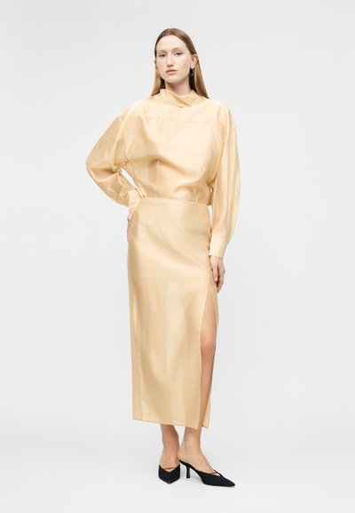 Γυναίκα που στέκεται με το ένα χέρι στη μέση, φοράει μπεζ μπλούζα με μακριά μανίκια και φούστα στο ίδιο χρώμα με ψηλή λωρίδα, μαύρες μυτερές γόβες, ουδέτερο φόντο.