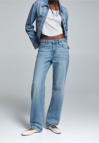 Mujer con chaqueta de mezclilla azul clara y jeans, camiseta corta blanca y bailarinas blancas, de pie frente a un fondo gris liso.
