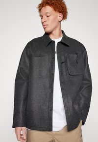 Veste en chemise noire en tissu tissé, dotée d'une poche avant zippée, d'une fermeture à bouton et d'une coupe décontractée.