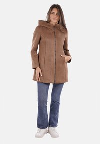 Cappotto in suede sintetico marrone con colletto alto, chiusura con zip e tasche laterali, abbinato a jeans a zampa d'elefante e sneakers bianche.