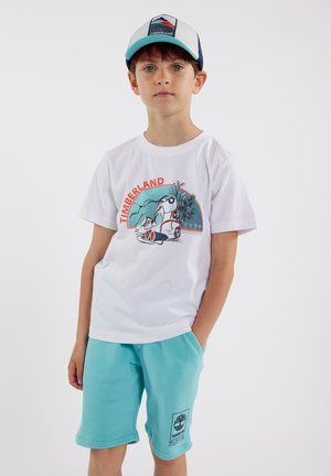Witte t-shirt met korte mouwen en kleurrijke "TIMBERLAND" grafiek en turkooise shorts met een logo-patch. Beide zijn gemaakt van zachte, lichte stof.